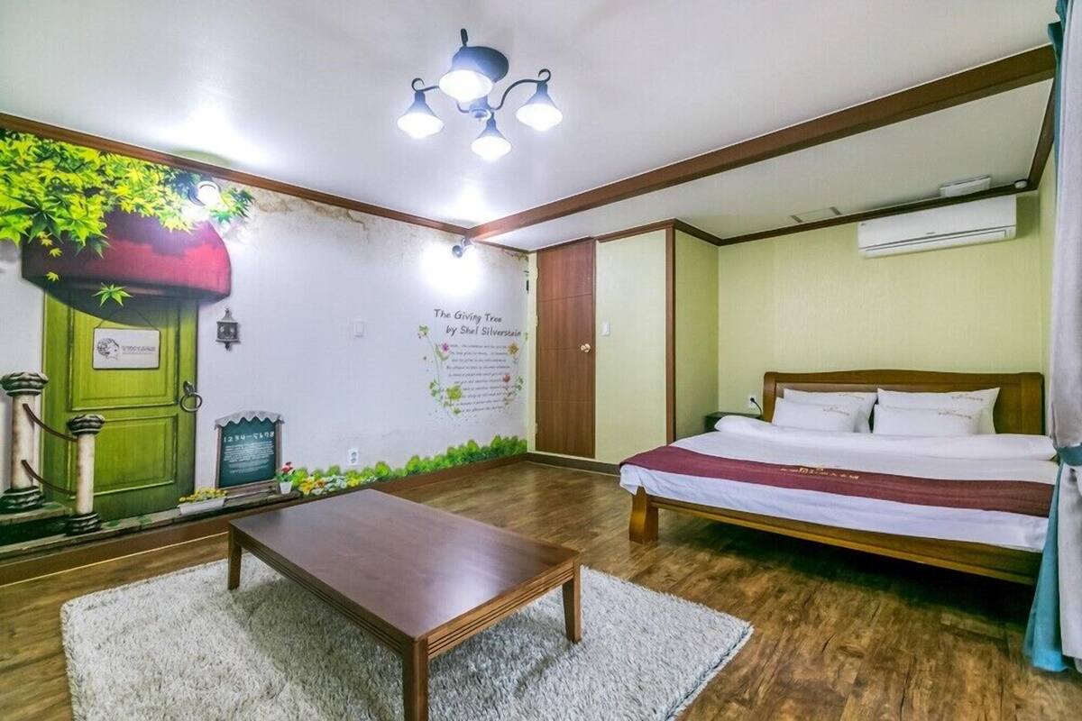 Una habitación con una cama, una mesa de madera, una puerta verde, un mural en la pared y una lámpara en el techo.
