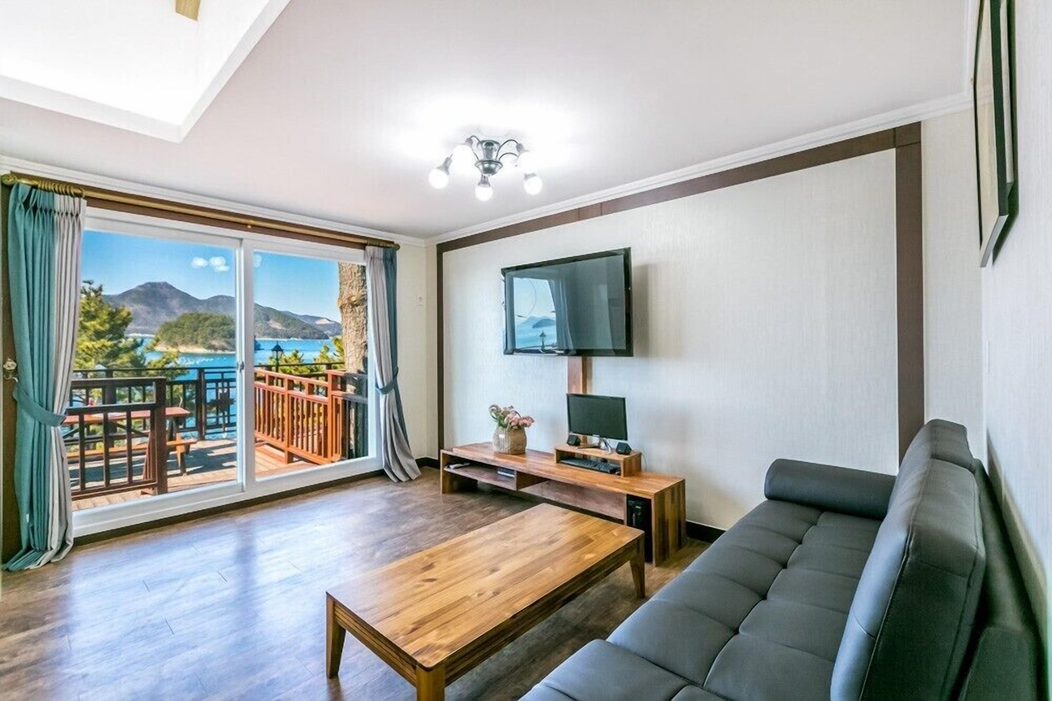Una sala de estar con un sofá, una mesa de centro de madera, un televisor sobre un mueble bajo y un balcón con vista al mar.