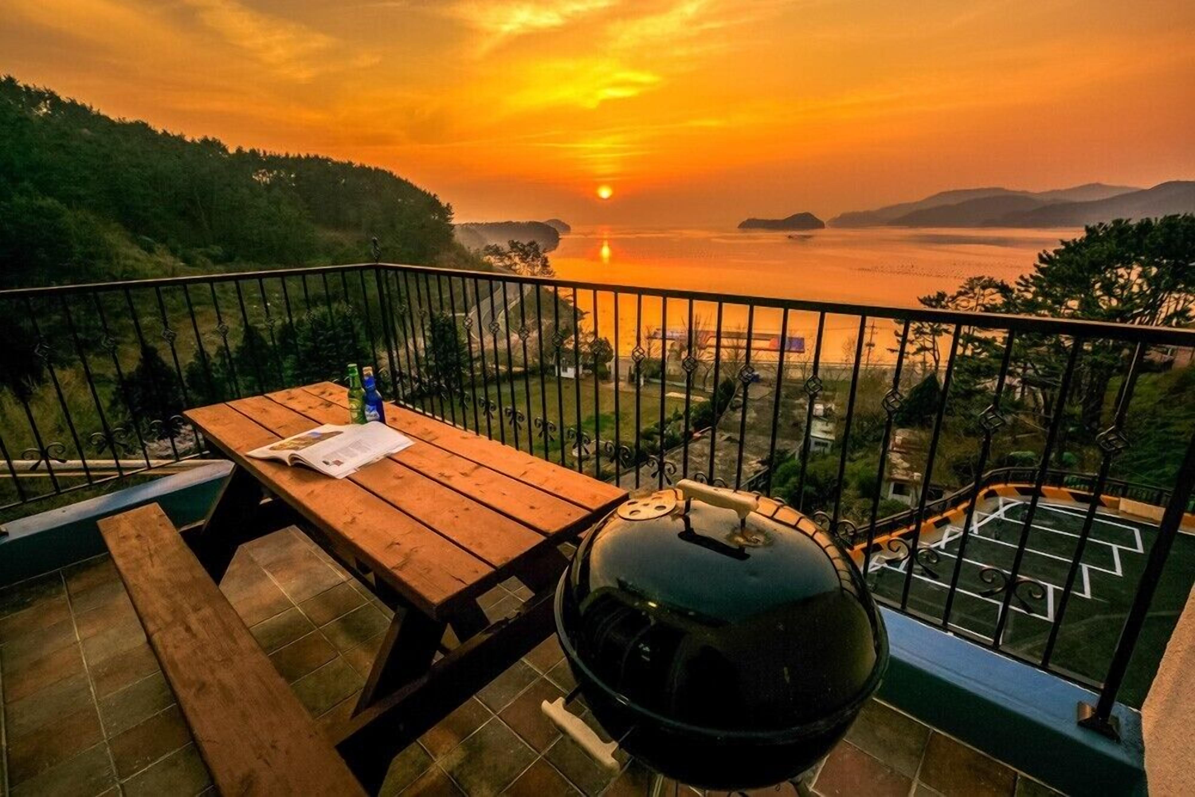 Un balcón con mesa de madera, una parrilla y vistas a un atardecer sobre el agua.