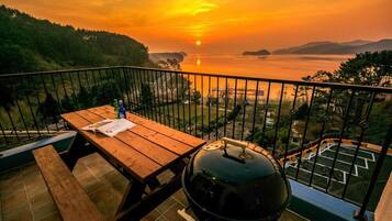 Un balcón con mesa de madera, una parrilla y vistas a un atardecer sobre el agua.