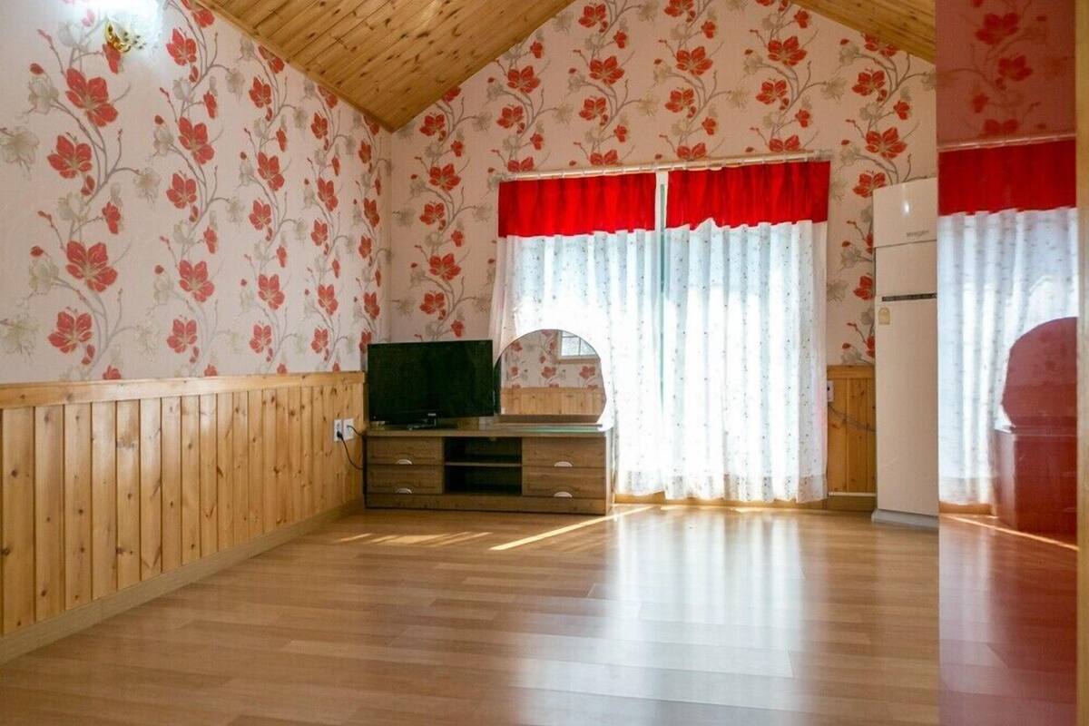 A room with wooden walls and floors, a wooden entertainment unit, a television, a window with red curtains, and a refrigerator.