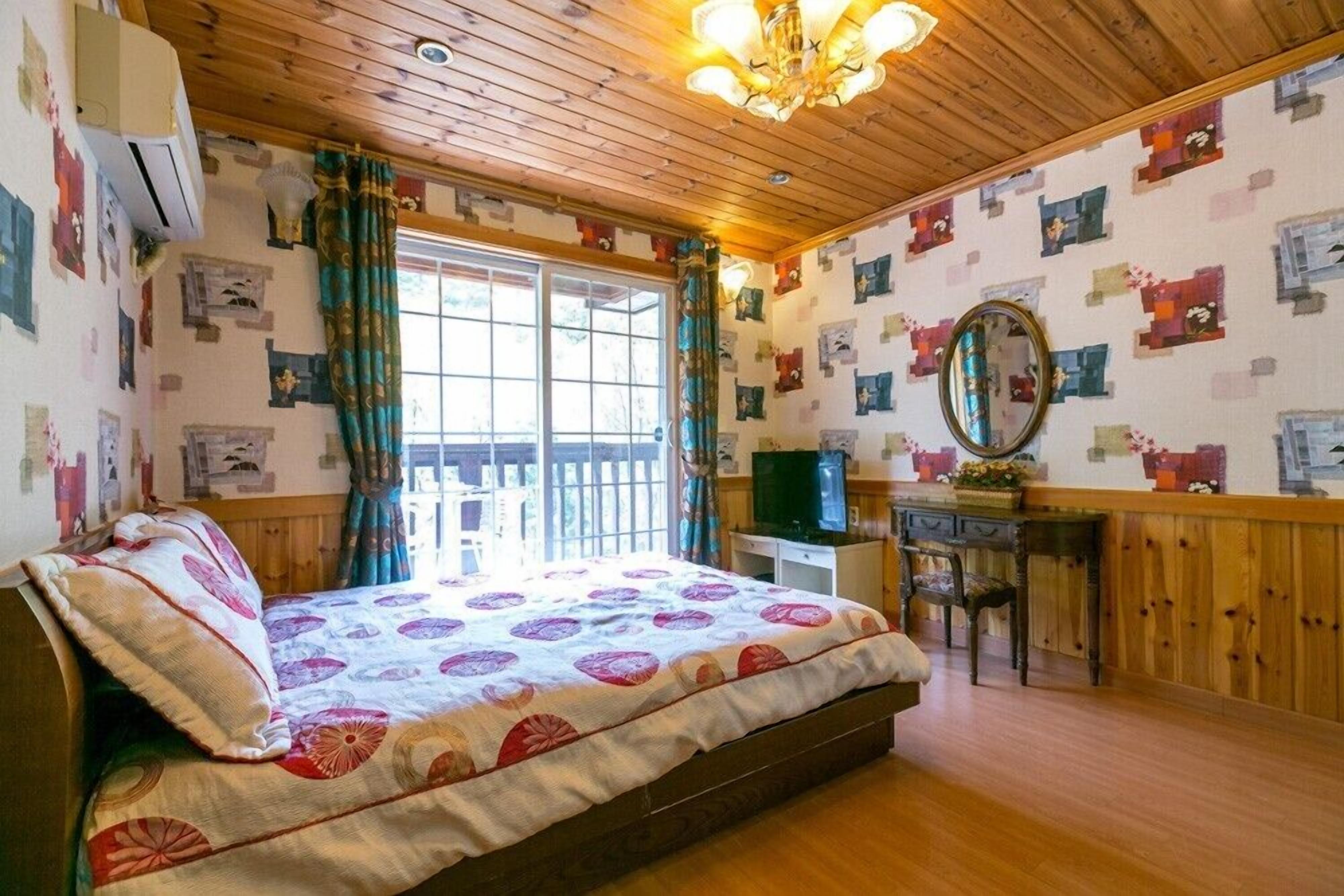 Un dormitorio con techo de madera, una cama con colcha estampada, un escritorio con espejo y una ventana con cortinas.