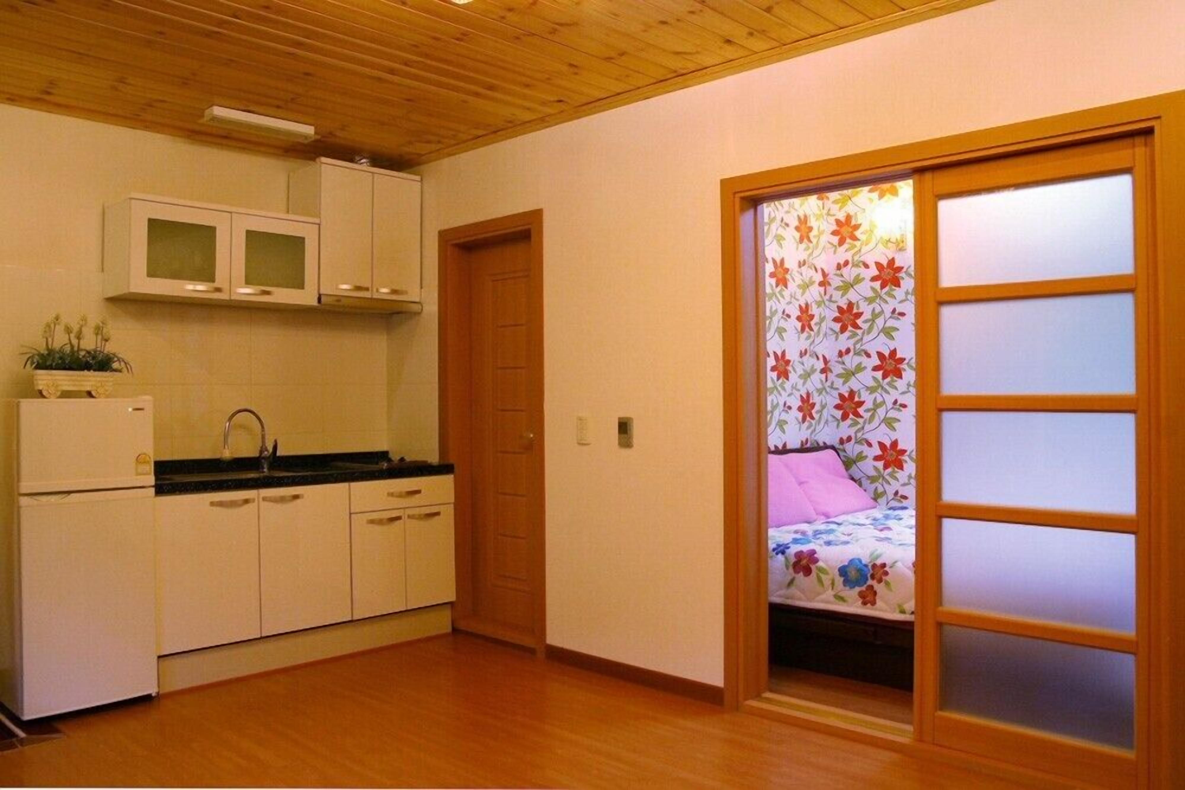 Une cuisine de taille réduite avec des meubles blancs et un plan de travail noir, une porte en bois et une porte-fenêtre coulissante donnant sur une chambre à coucher décorée de rideaux à motifs floraux.