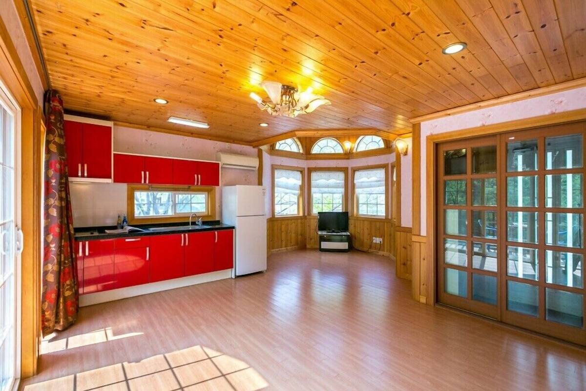 A spacious room with wooden flooring, a kitchen with red cabinets, and large windows offering views of the outdoors.