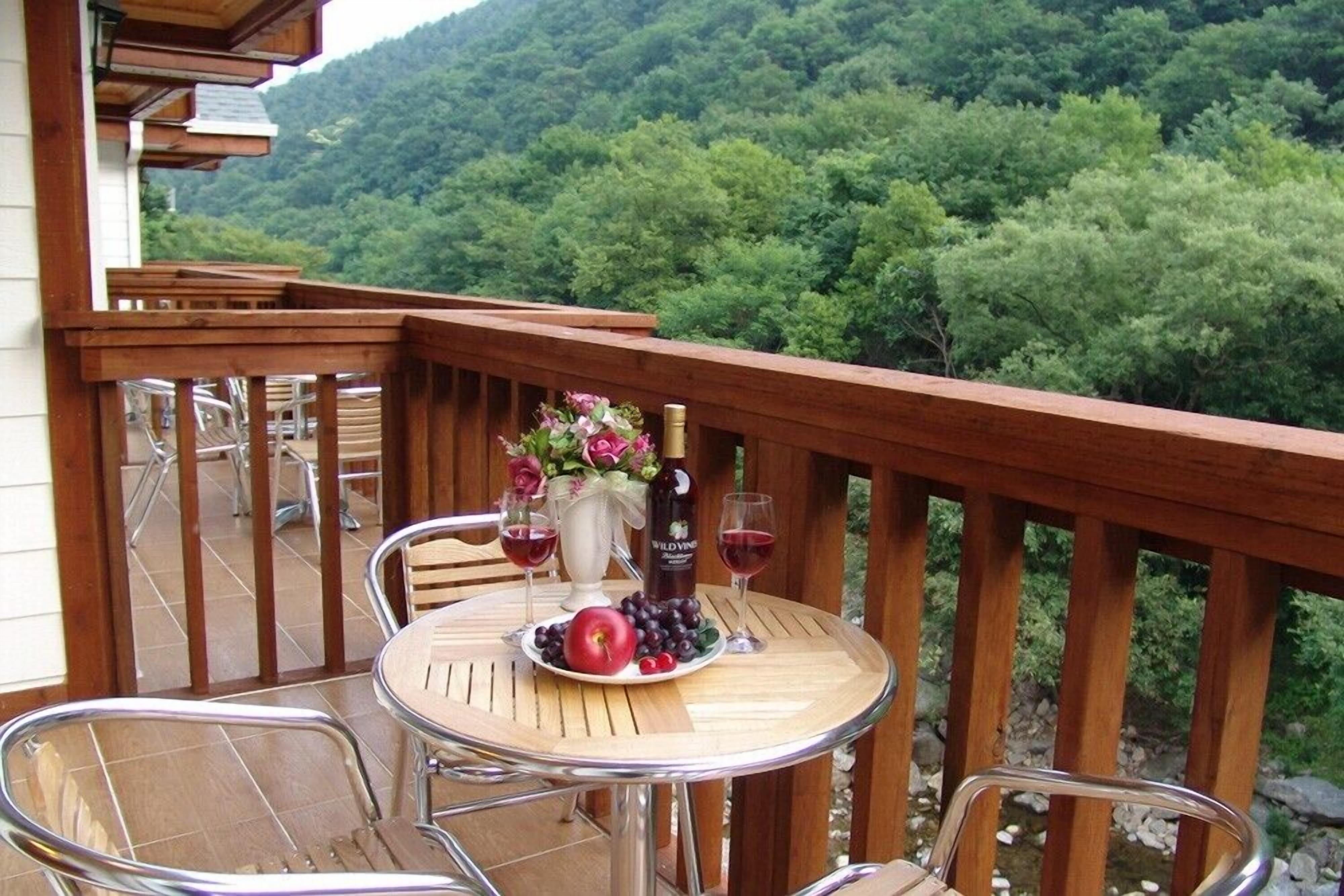 A wooden deck with a round table set for two, featuring a bottle of wine, glasses, and a plate with grapes and an apple.