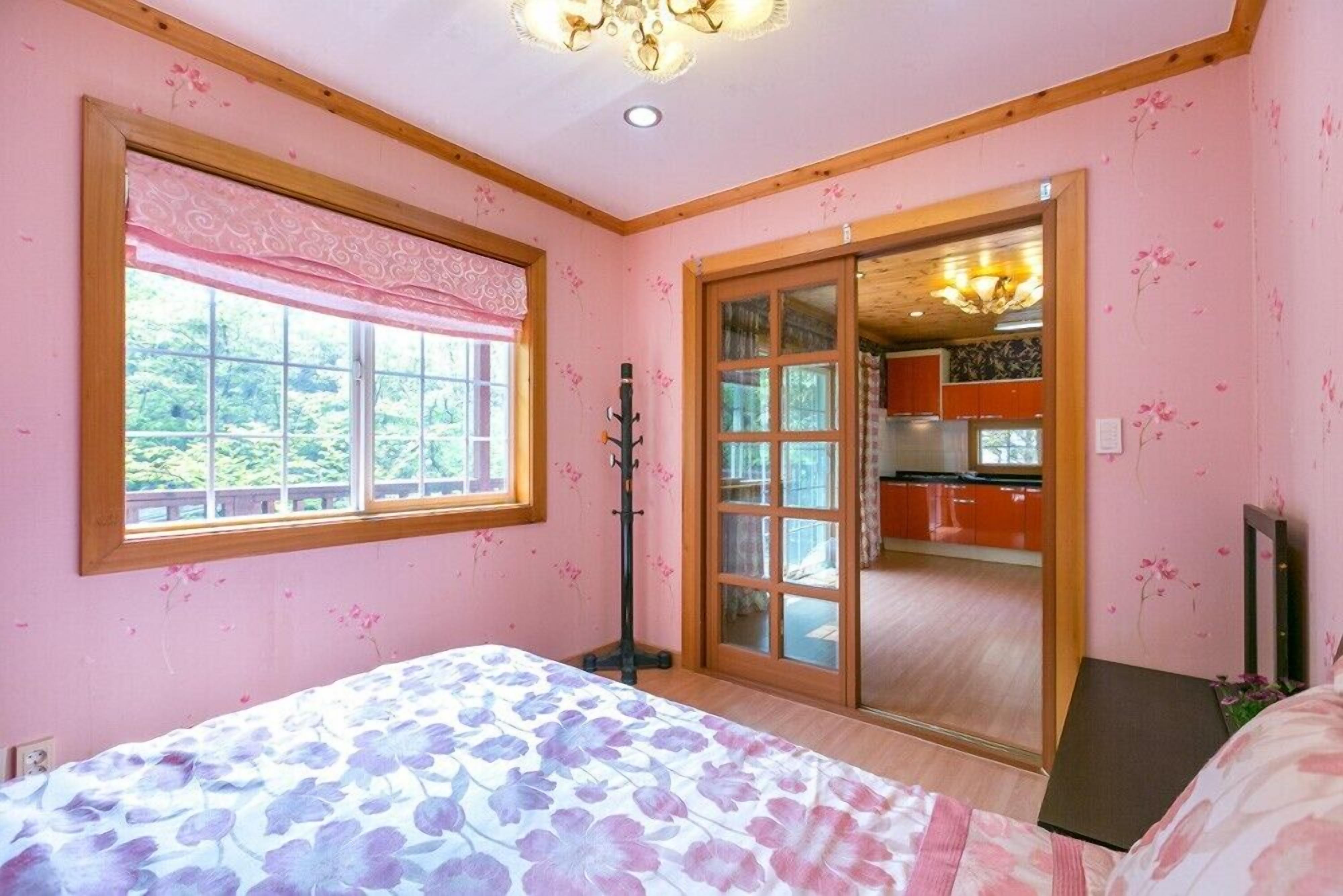 A bedroom with a bed, a window, a door leading to a kitchen, and a ceiling light.