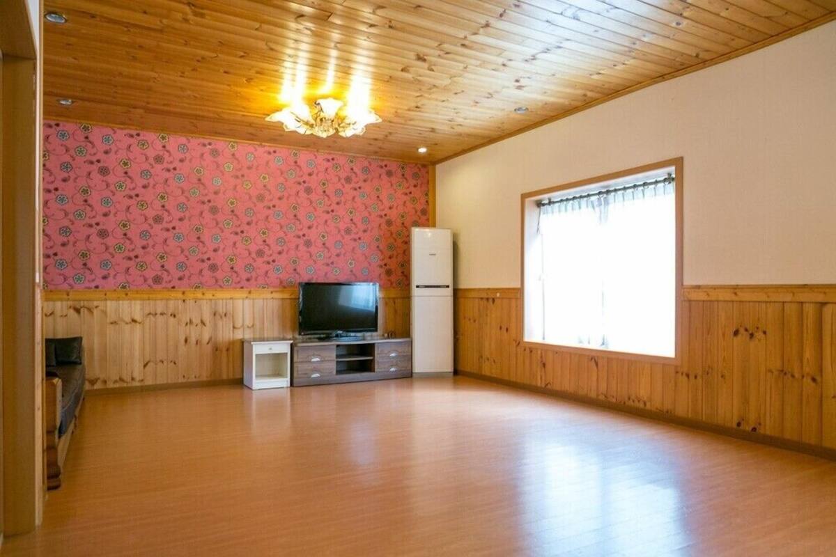 A spacious room with wooden flooring, a television on a wooden stand, a refrigerator, and a window with curtains.