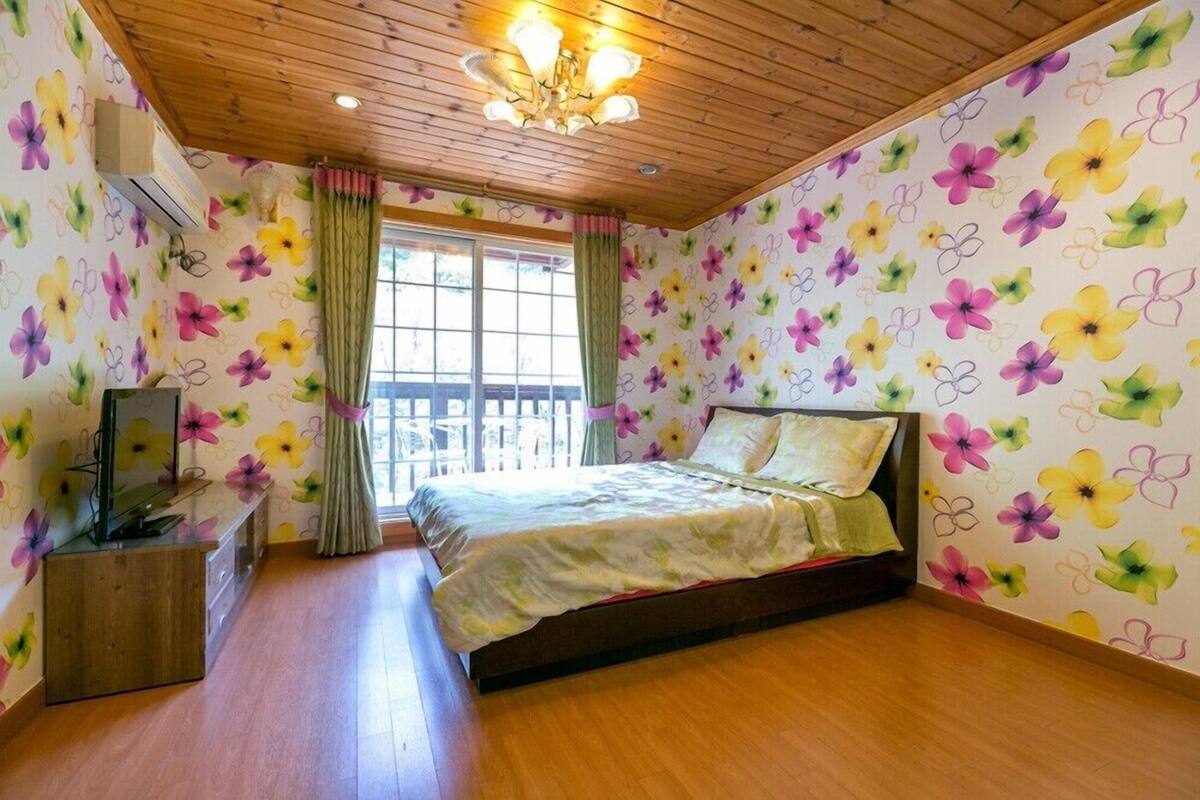 A bedroom with a wooden ceiling, a bed with a floral-patterned bedspread, a wooden dresser, and a window with curtains.