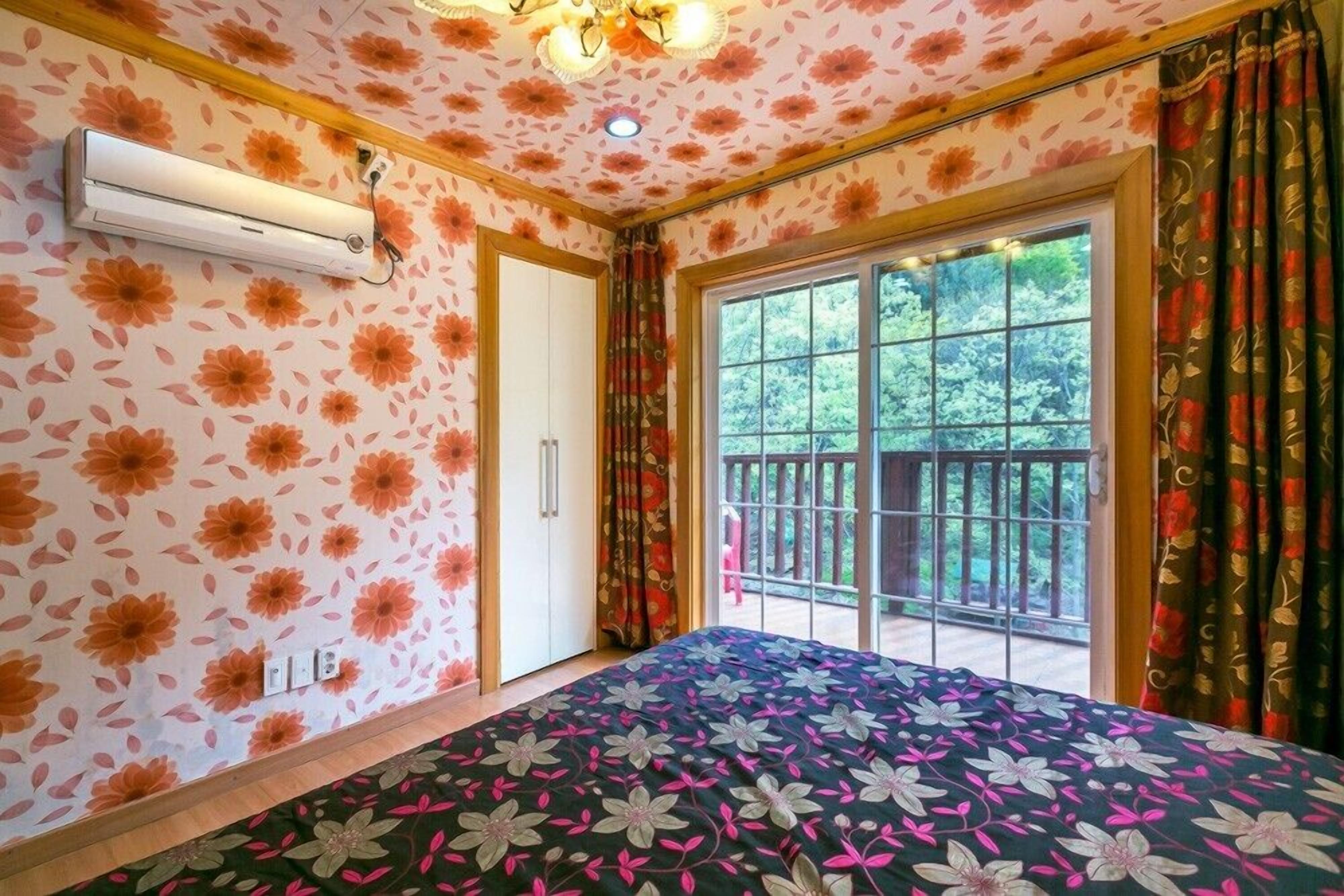 Una habitación con un papel tapiz estampado con motivos florales, una cama con colcha estampada, un ventanal corredero a un balcón y un equipo de aire acondicionado en la pared.