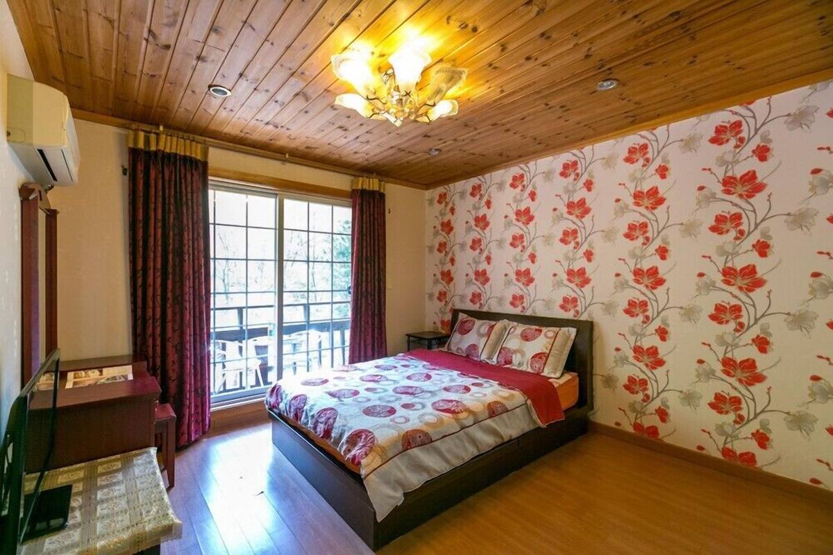 A bedroom with a wooden ceiling, a bed with floral bedding, a desk, and a window with curtains.