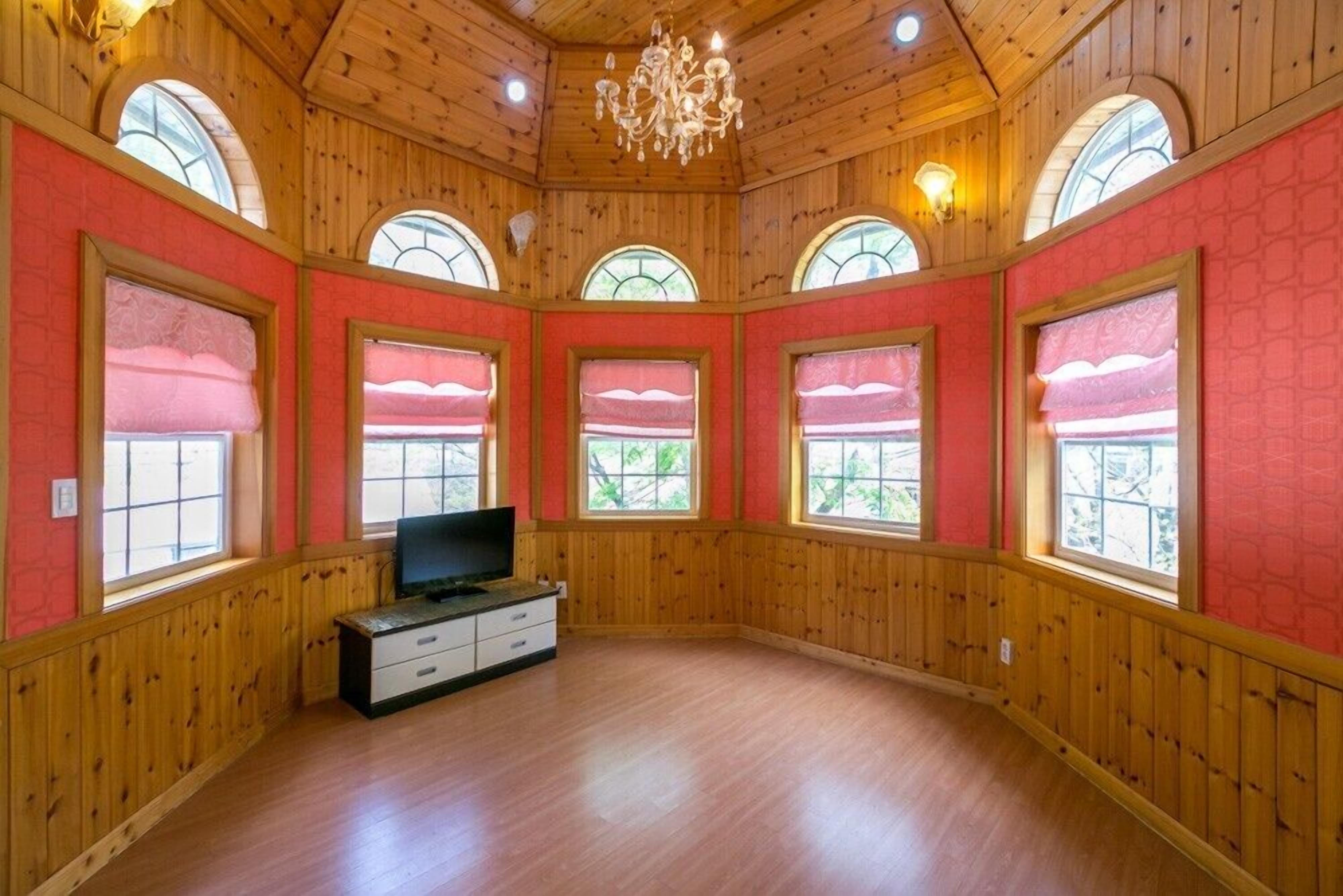Una habitación con paredes revestidas de madera, ventanas arqueadas grandes con cortinas rojas, una araña y un televisor de pantalla plana sobre un aparador de madera.