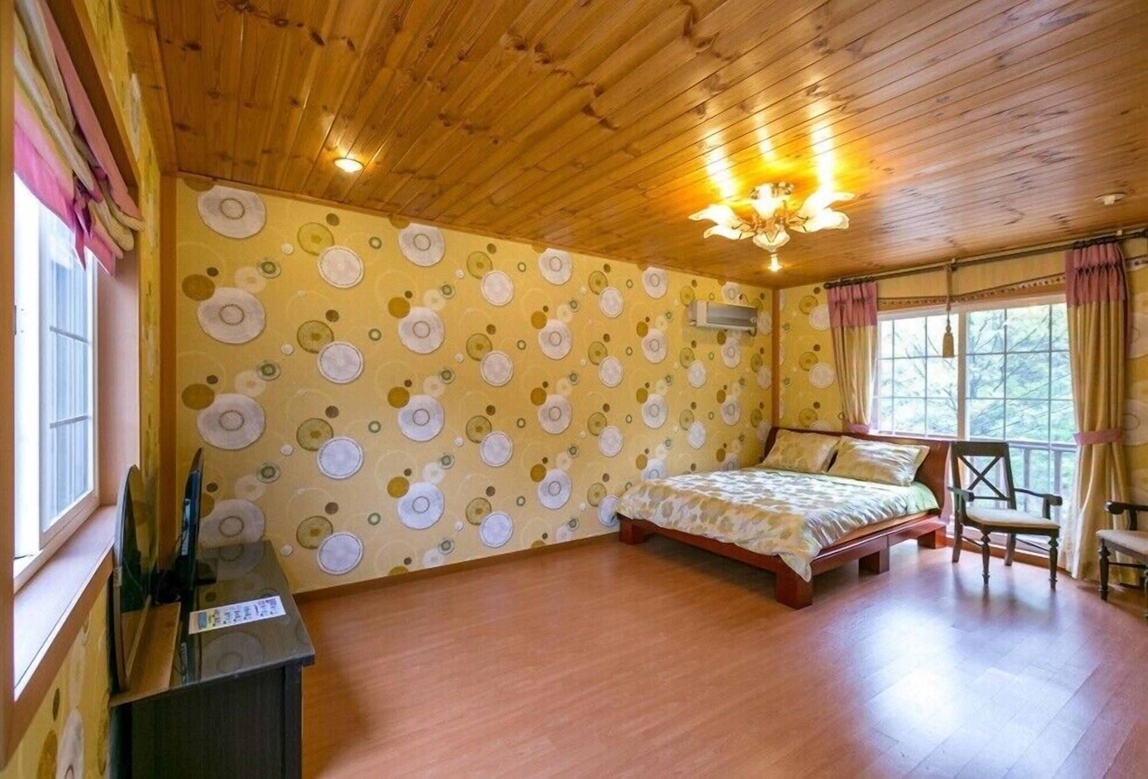 A room with a wooden ceiling, a bed with a patterned bedspread, a chair, and a window with curtains.