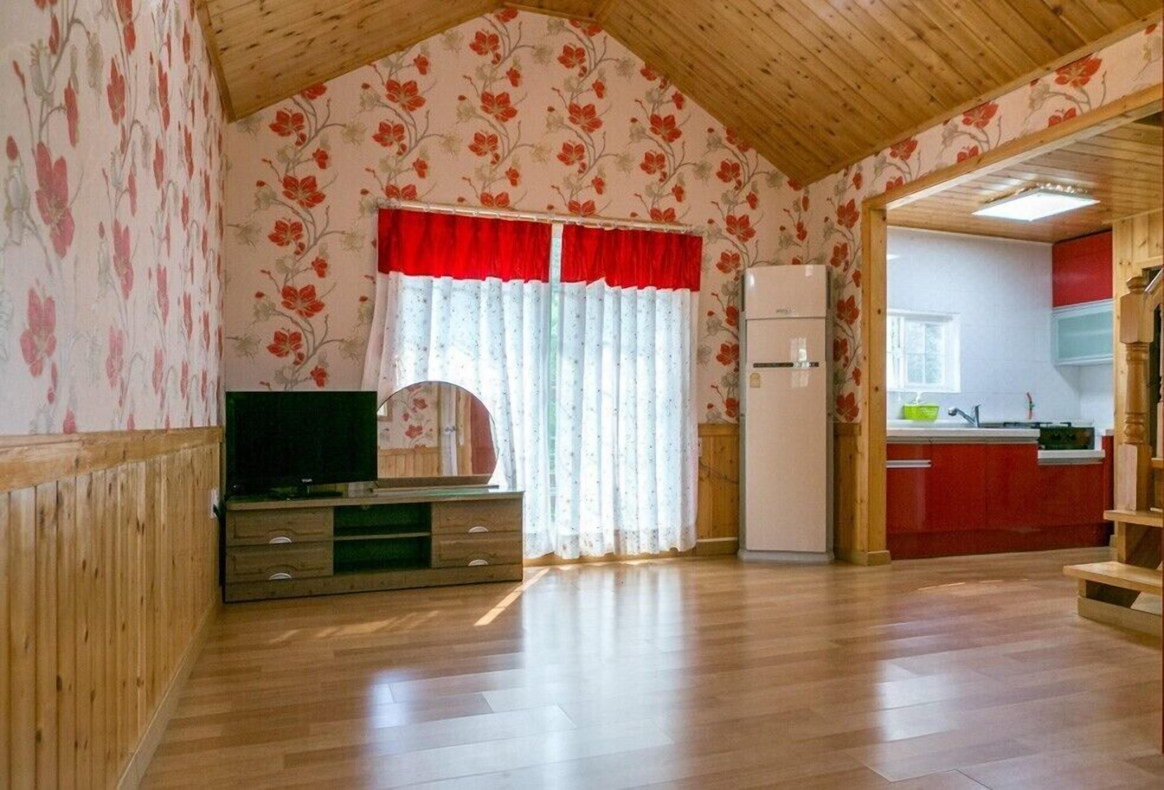 A room with wooden flooring, a television on a wooden cabinet, a refrigerator, and a kitchen area with red cabinets and a window.