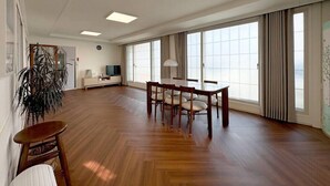 A spacious living room with a wooden floor, a dining table with chairs, a TV, and large windows with curtains. - Taeyoung Pension (Pohang)
