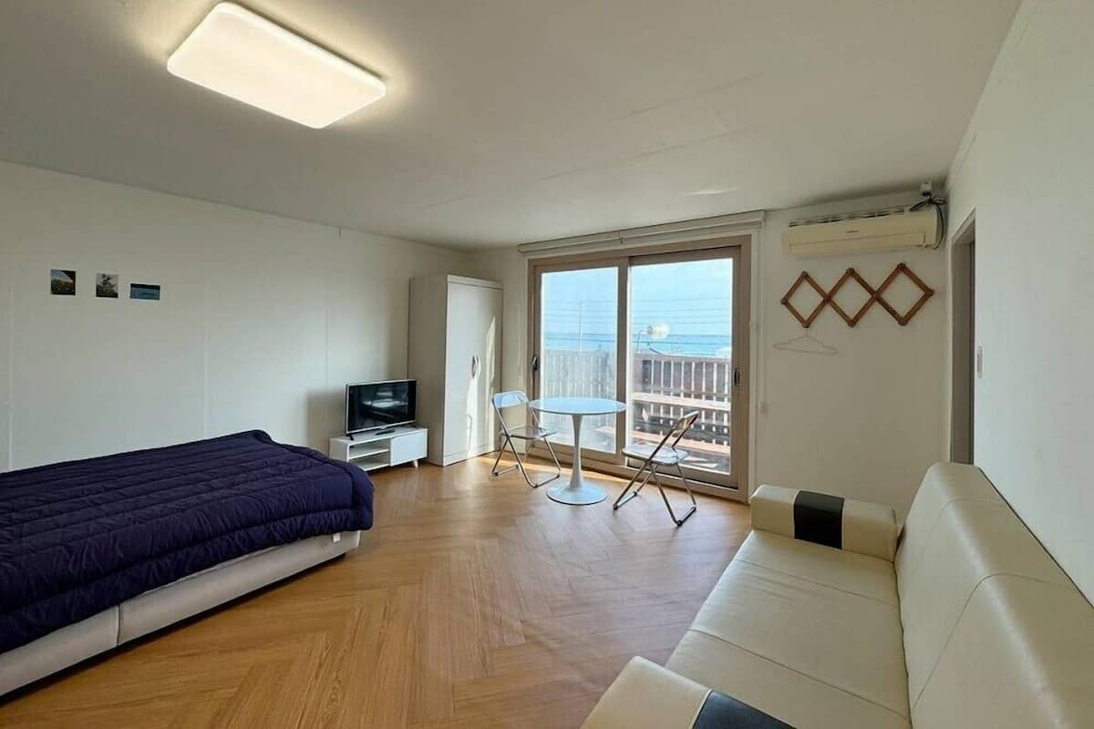 A modern hotel room with a bed, a sofa, a small table, and chairs. There is a TV on a stand, a window with a view, and an air conditioning unit on the wall.