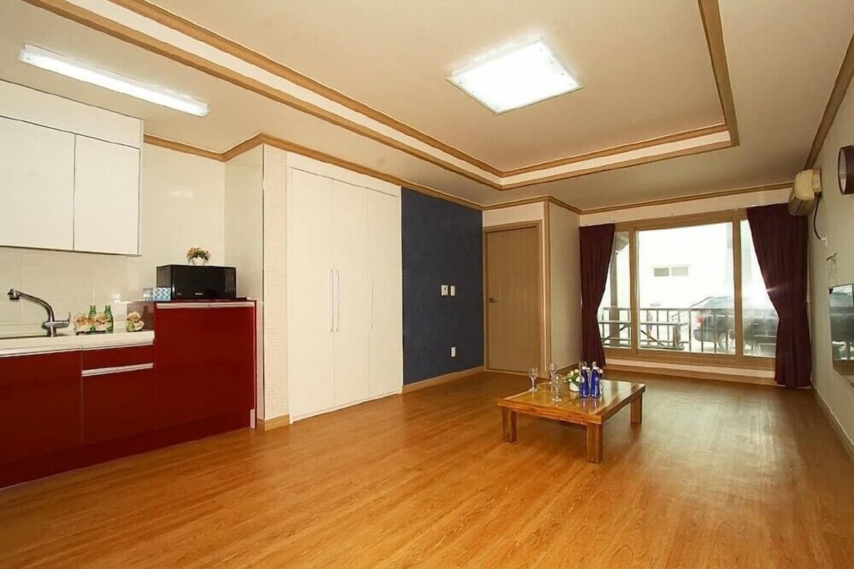 A spacious living area with a wooden floor, a kitchen with red cabinets, a dining table with wine glasses, and a large window with curtains.