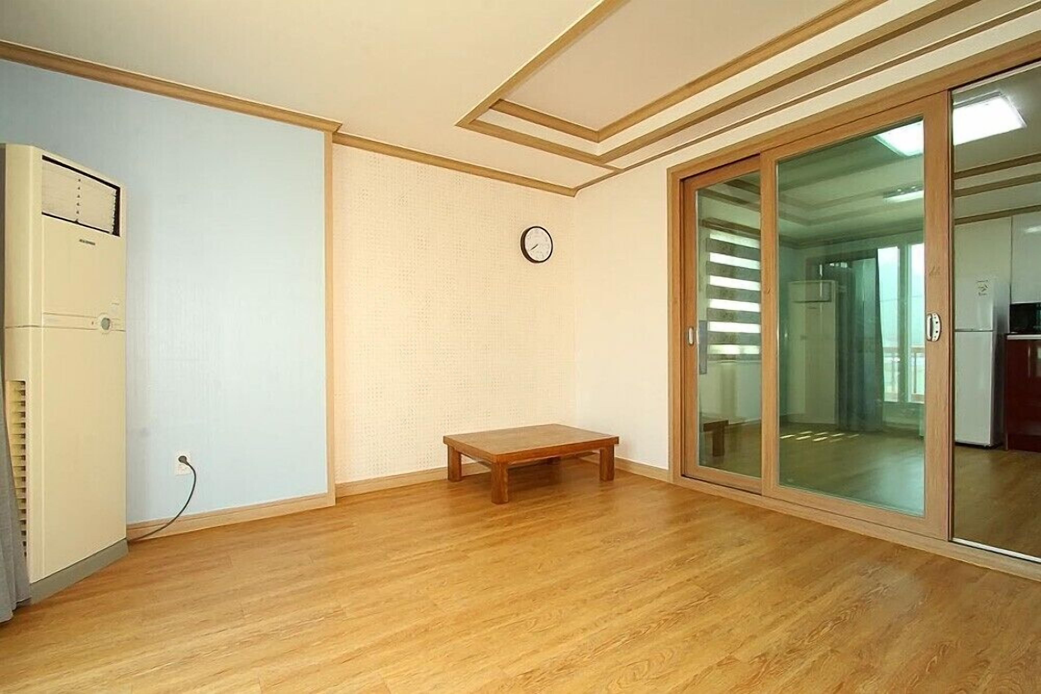 A room with a wooden floor, a small wooden bench, a white air conditioning unit, and a sliding glass door leading to another room.