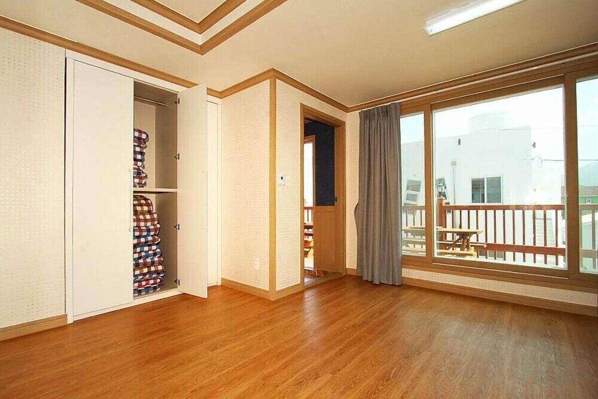 A spacious room with wooden flooring, a large window with a balcony, and a wardrobe with folded clothes.