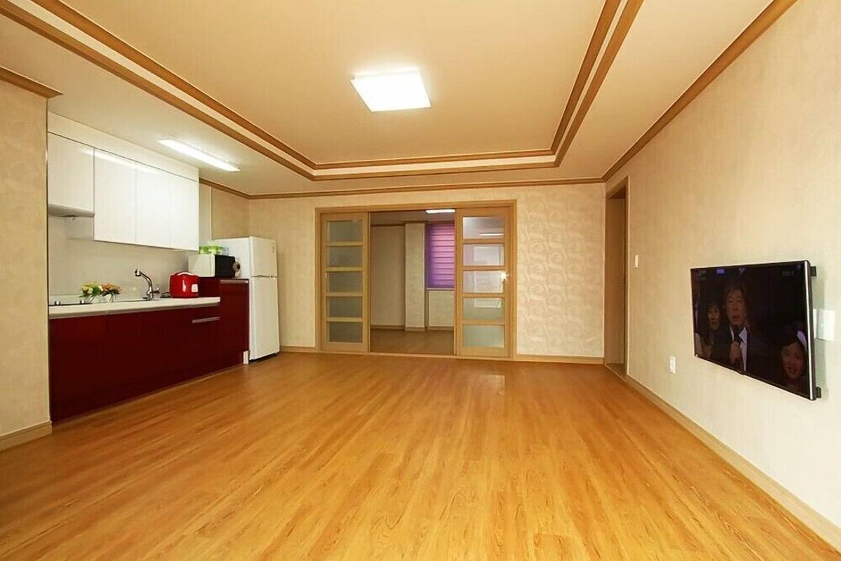 A spacious room with a wooden floor, a kitchen area with white cabinets, and a door leading to another room.