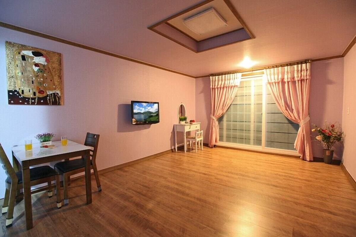 A room with a wooden floor, a dining table with chairs, a TV, a window with curtains, and a painting on the wall.