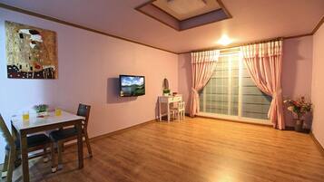 A room with a wooden floor, a dining table with chairs, a TV, a window with curtains, and a painting on the wall.