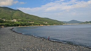 Una spiaggia con ciottoli e un mare calmo, colline in lontananza e alcuni edifici sullo sfondo.
