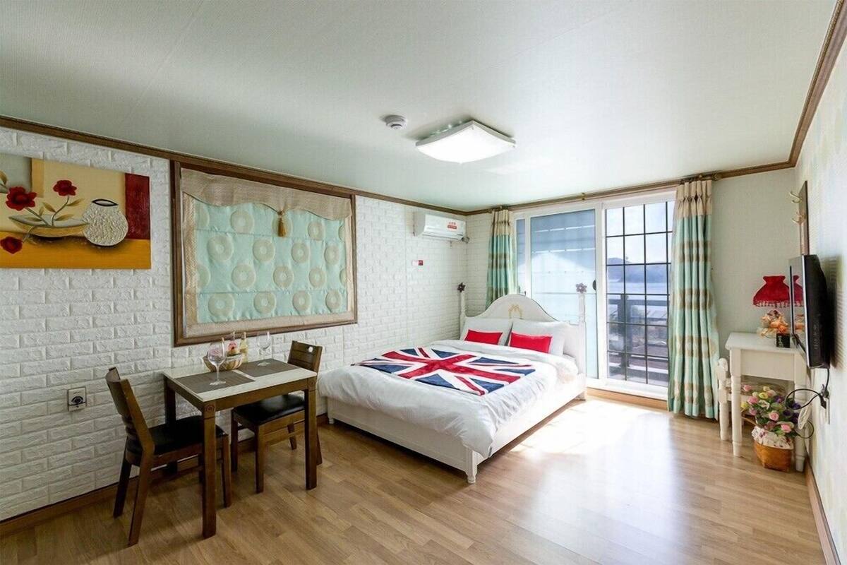 A bedroom with a bed, a desk, and a chair. There is a window with curtains and a view outside.