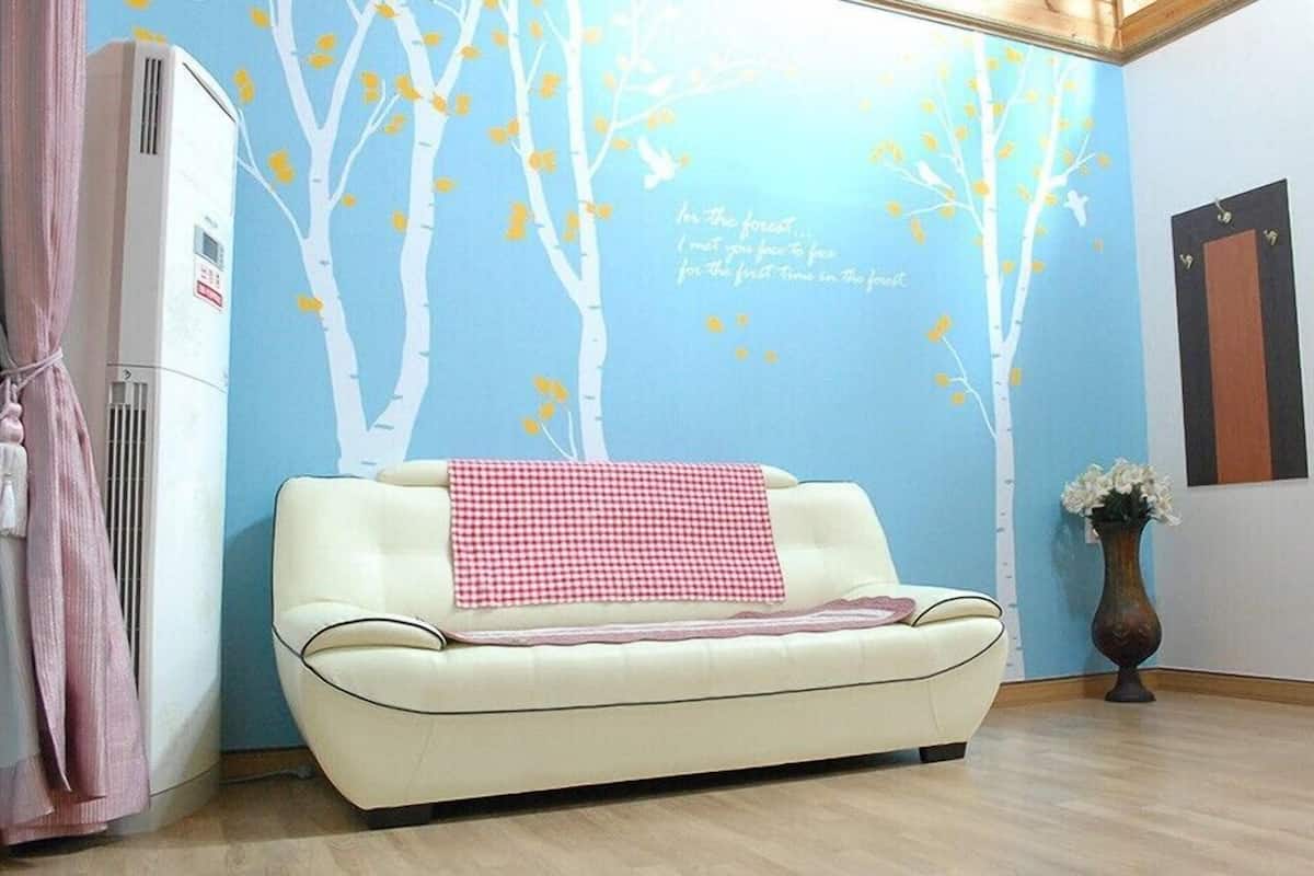 A room with a white sofa, a wall mural of trees, a small vase with flowers, and a radiator.