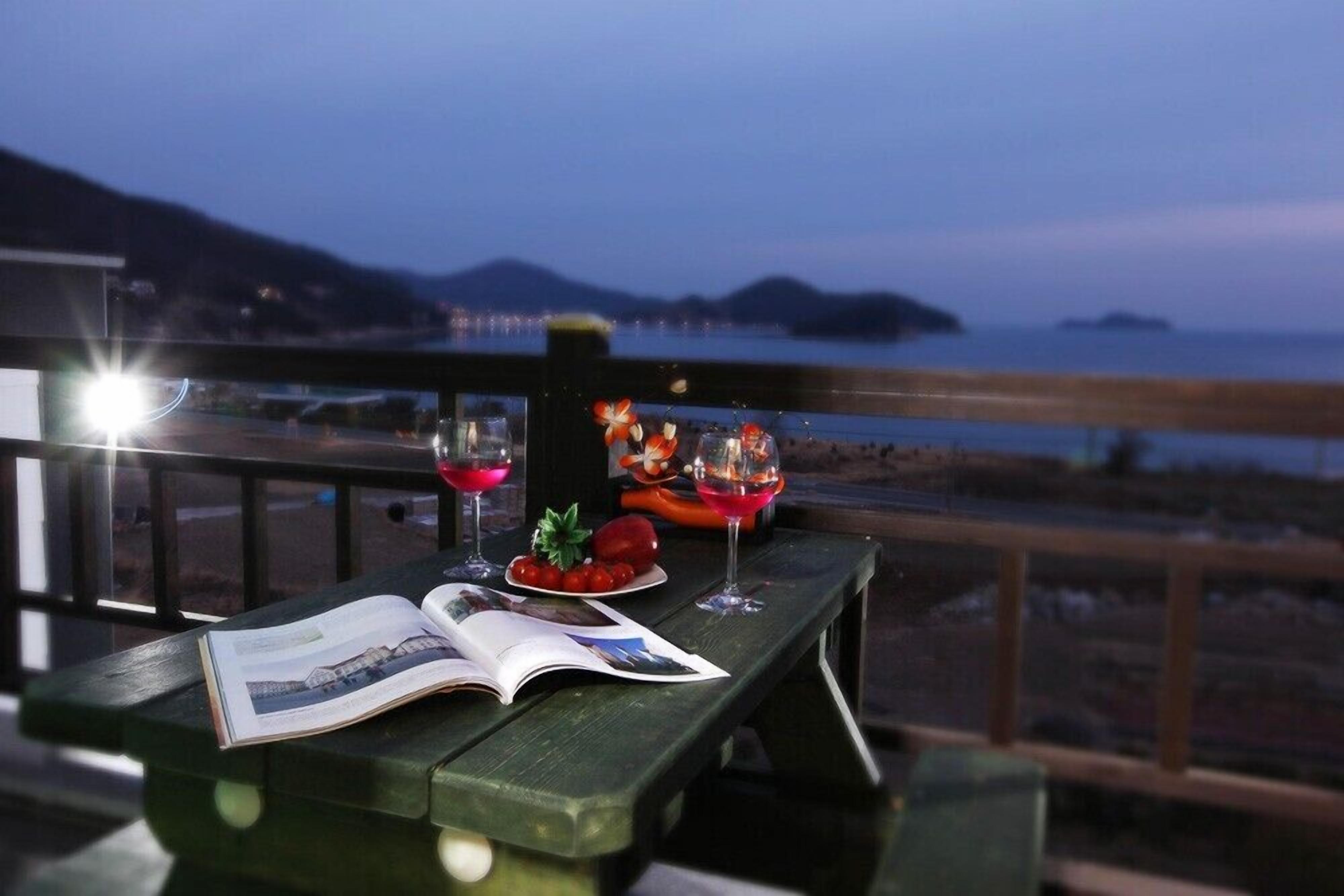 Un banc de fusta amb un llibre i un parell de copes de vi tinto, amb vista a una zona costanera i muntanyes llunyanes, a més d'una massa d'aigua.