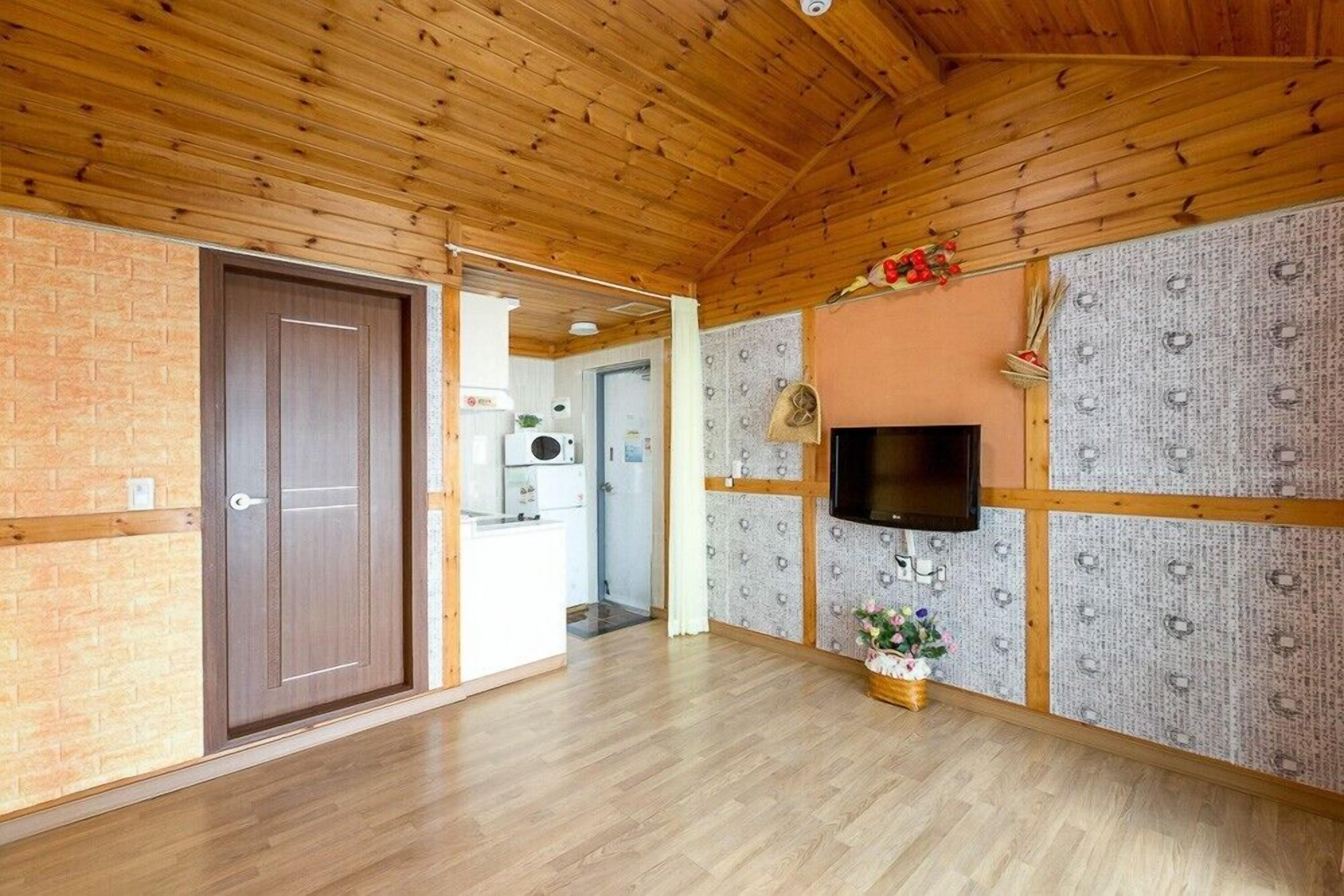 A spacious room with a wooden ceiling and walls, a flat-screen TV mounted on the wall, and a small kitchen area in the background.