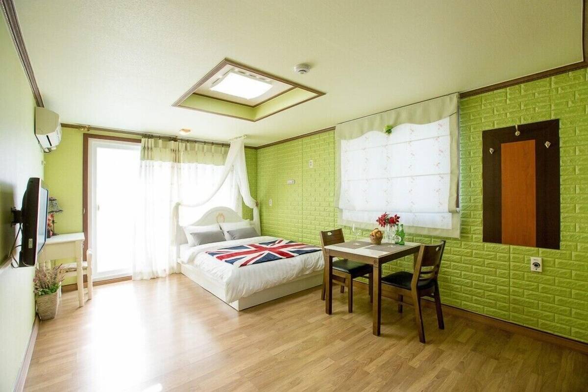 A room with a bed, a table with chairs, a window with curtains, a wall-mounted TV, and a door.