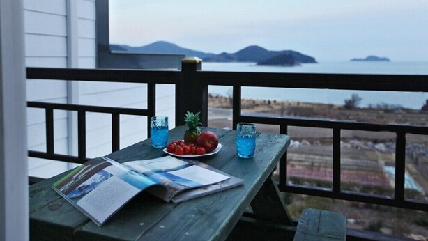 Un tavolo di legno con una rivista, un piatto di frutta e bicchieri di bevanda blu su un balcone affacciato sull'acqua e sulla costa in lontananza.