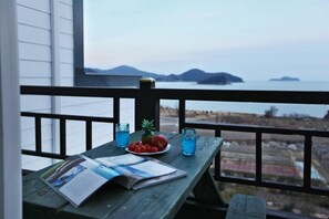 Ein Holztisch mit einer Zeitschrift, einer Fruchtschale und Gläsern mit blauem Getränk auf einem Balkon mit Blick auf ein Gewässer und entfernte Küsten.