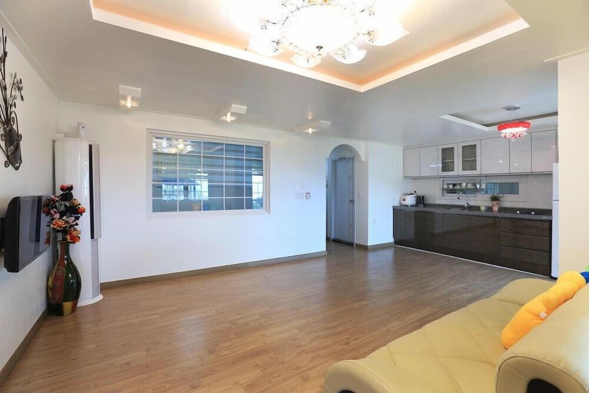A spacious living area with a wooden floor, a sofa, a kitchen with dark cabinets, and a large window with blinds.