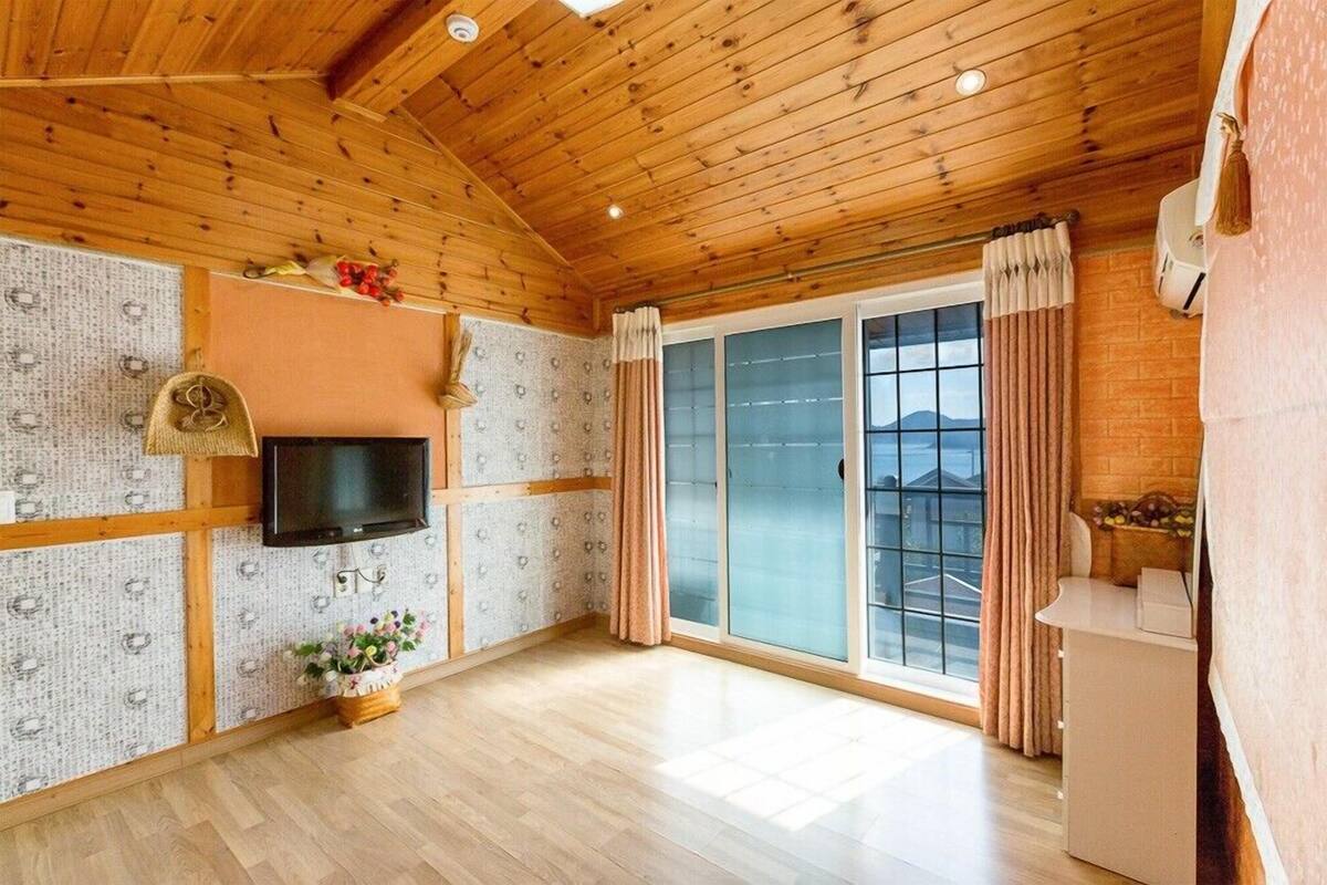 A spacious room with wooden ceiling beams, a flat-screen TV mounted on the wall, a sliding glass door leading to a balcony, and a small kitchenette area.