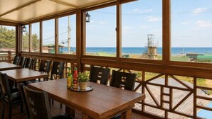 Un espacio de comedor al aire libre cubierto, con mesas y sillas, grandes ventanales con vista al mar y una planta eléctrica.
