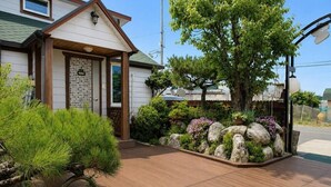 Una terraza de madera con borde de piedra y plantas, una casa con techo inclinado y un árbol grande al fondo.