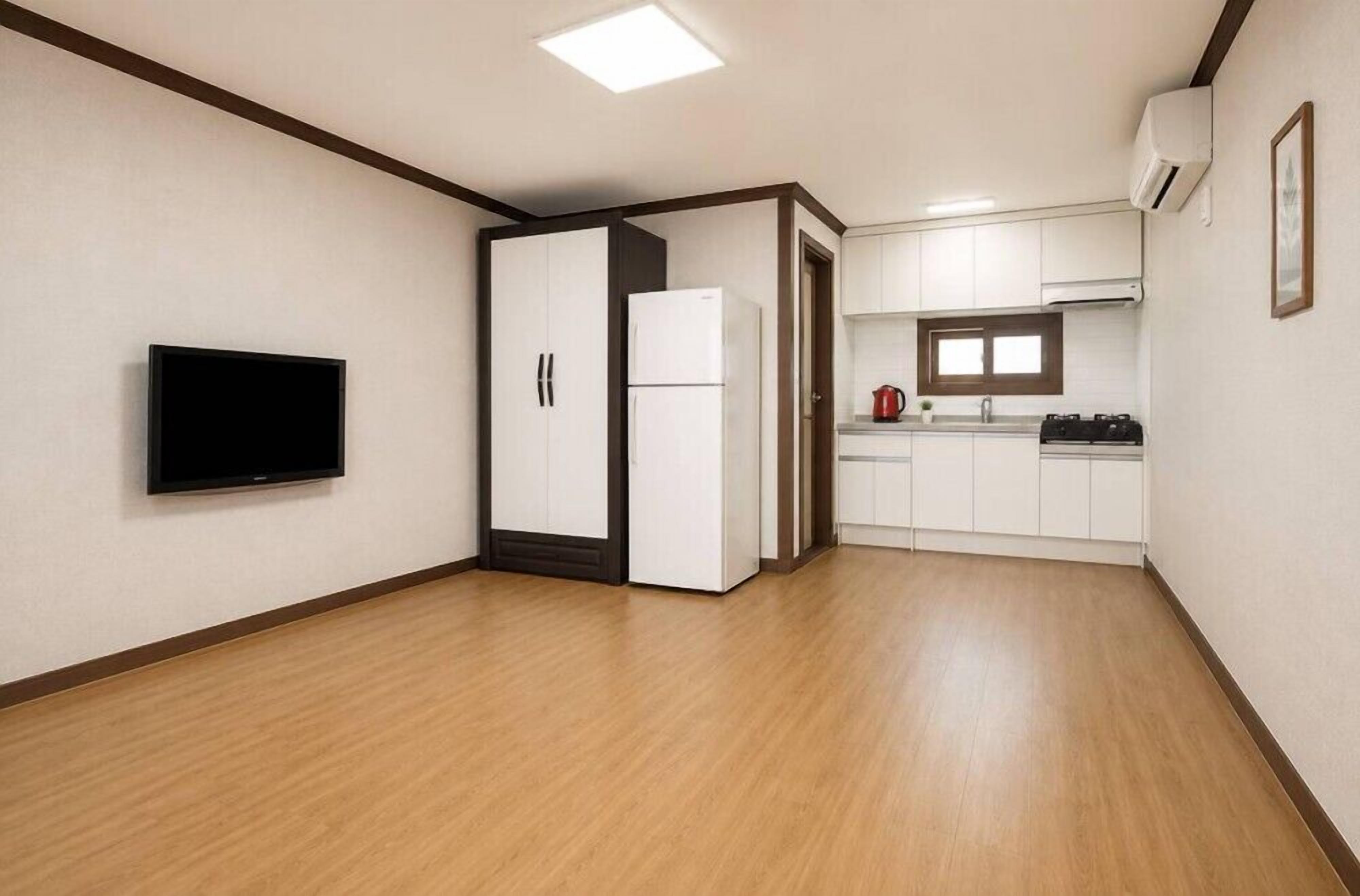 A room with a flat-screen TV, a white wardrobe, a white refrigerator, a kitchen area with cabinets, and a window.