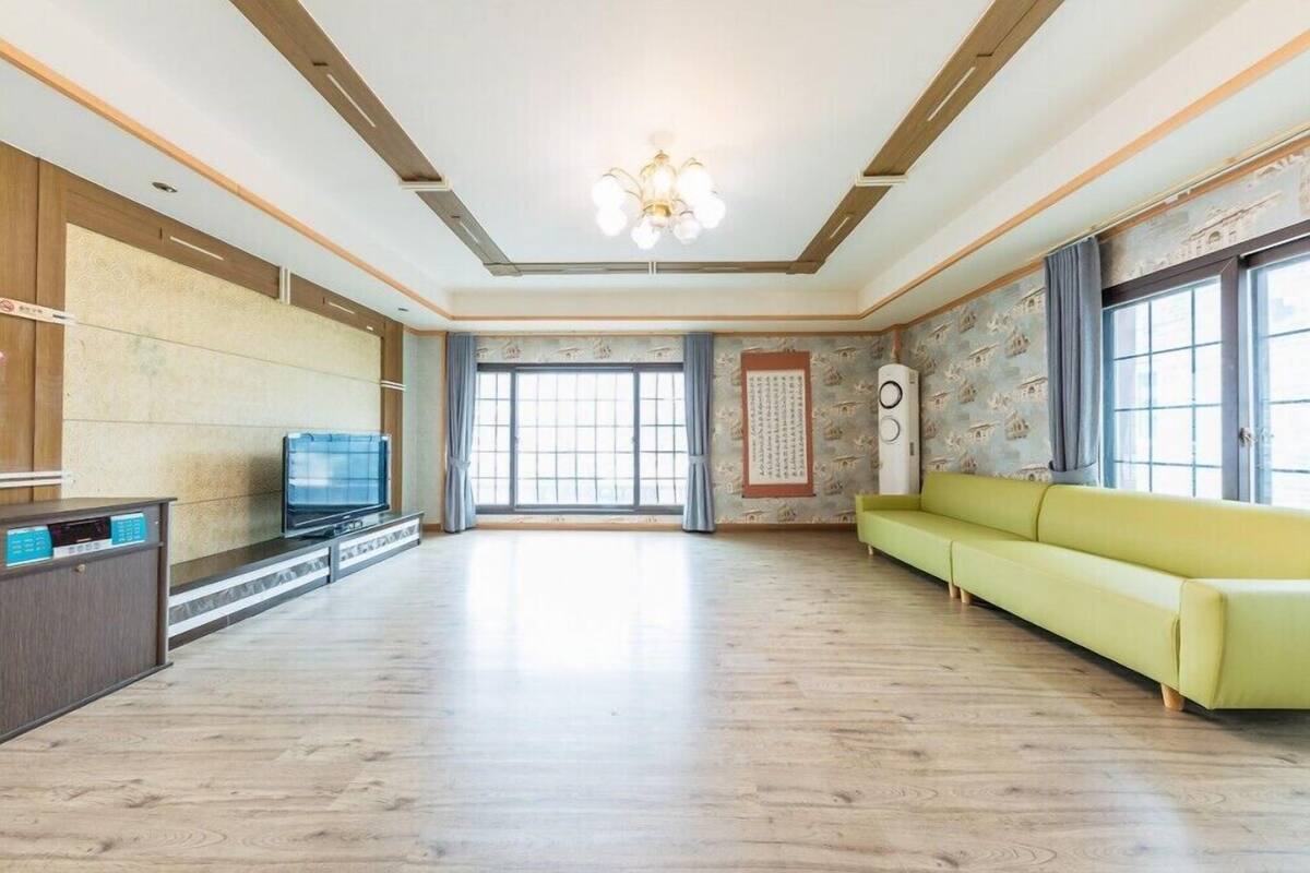A spacious living room with a wooden floor, a television, a sofa, and a large window with curtains.
