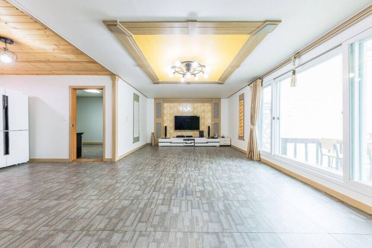 A spacious living room with a large TV, wooden ceiling, and large windows.
