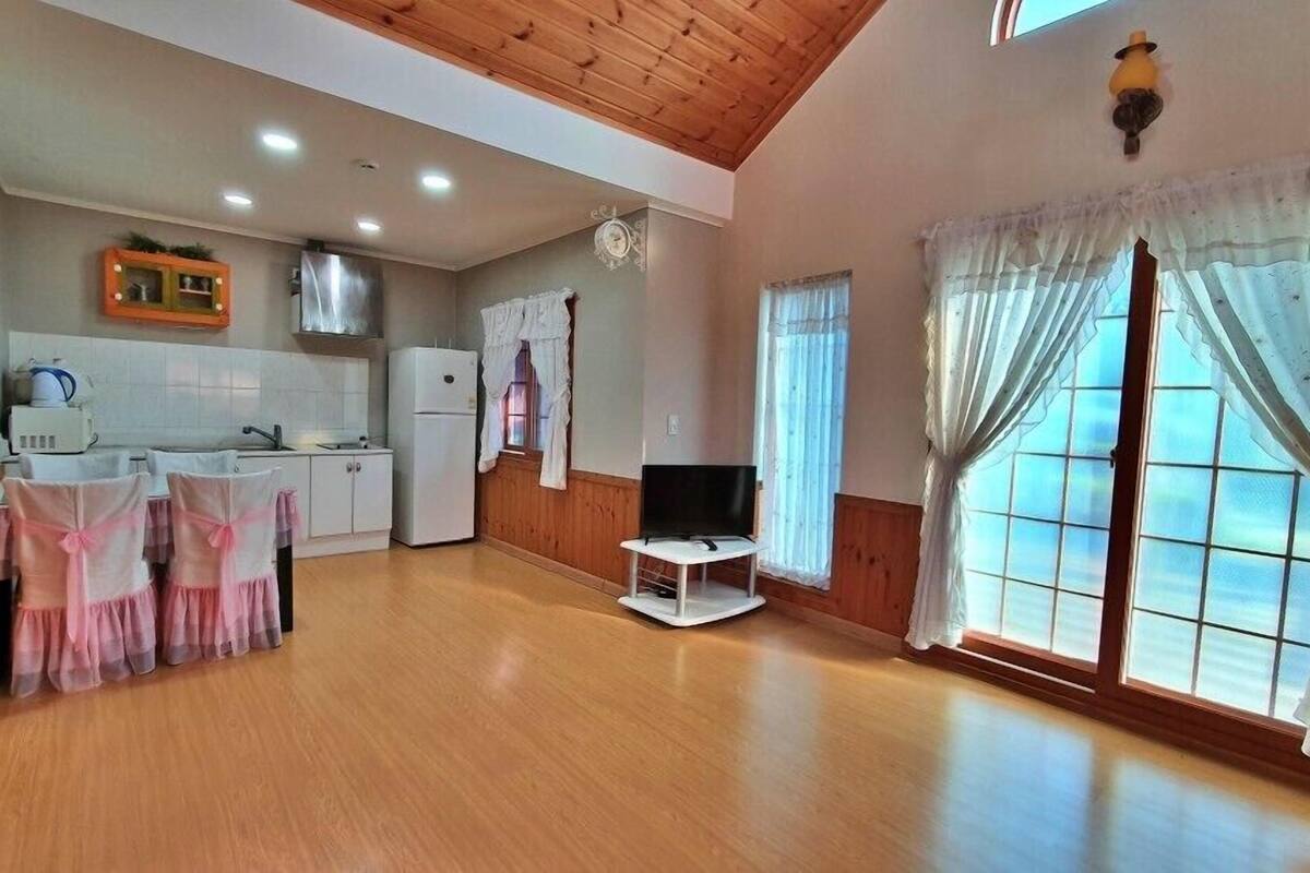 Ein großzügiger Wohnbereich mit Küche, Holzböden, Fernseher und großen Fenstern mit Vorhängen.