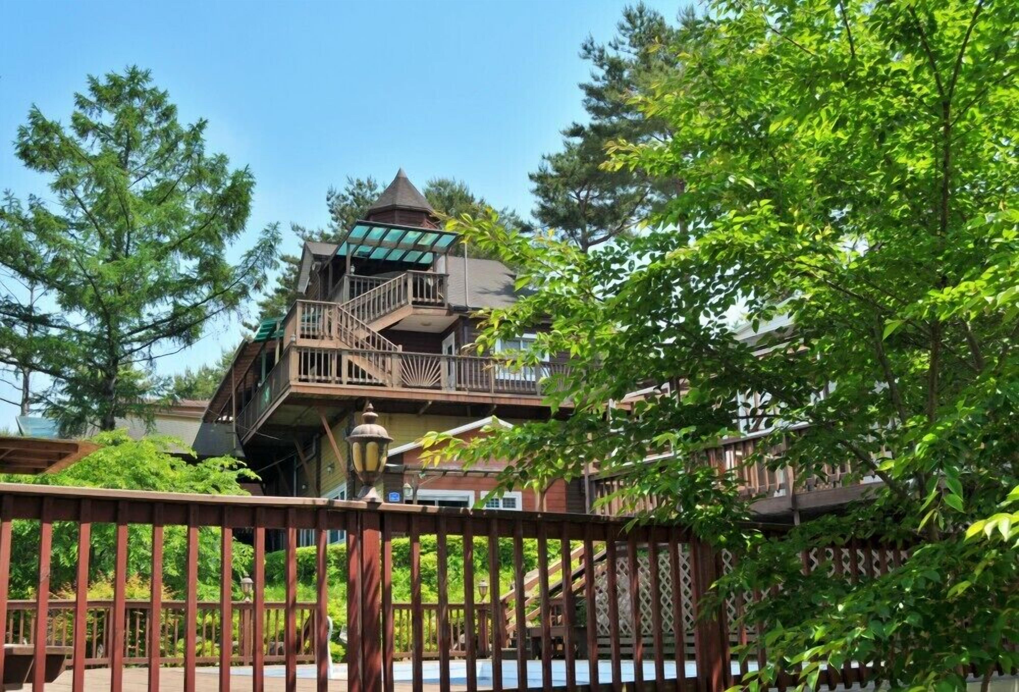 A multi-level wooden building with a balcony, surrounded by trees and a wooden fence.