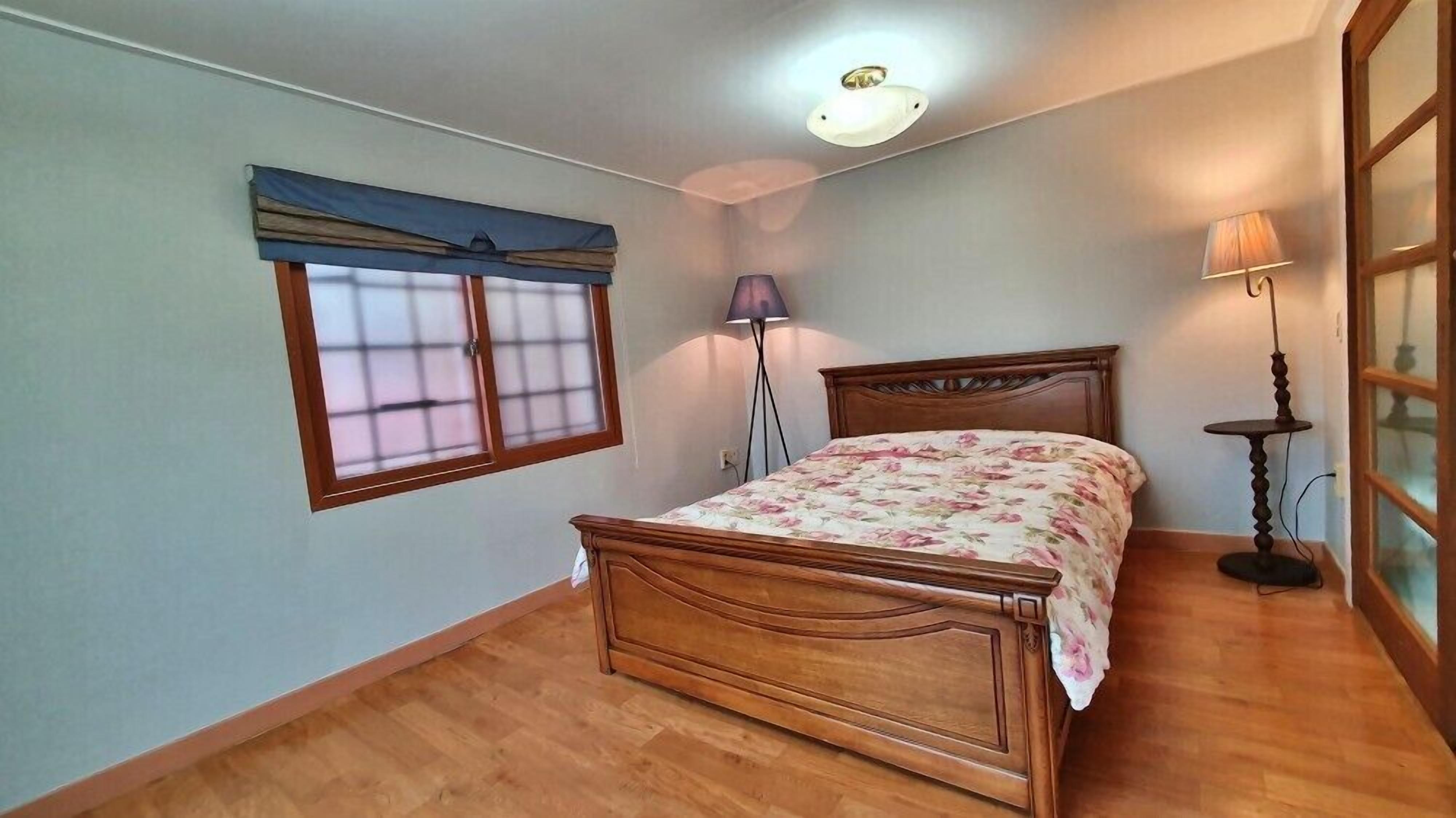 Un dormitorio con una cama de madera, una ventana con persianas, una lámpara de pie y una luz de techo.