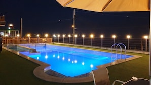 Una piscina al aire libre de noche con luces circundantes, una zona cubierta y algunas sillas de descanso.