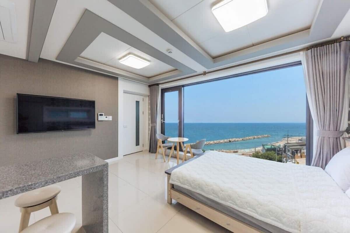 A modern hotel room with a large bed, a TV mounted on the wall, a small dining area, and a balcony with a sea view.
