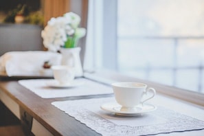 A table with a cup and saucer on a lace placemat, a vase with white flowers, and a white cloth on a wooden table near a window. - The Foret Pension (Gapyeong)