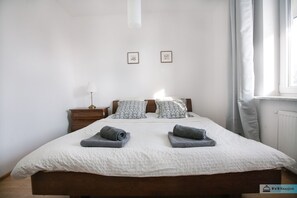 Ein ordentlich bezogenes Bett mit zwei aufgerollten Handtüchern, einem Nachttisch mit Lampe und zwei gerahmten Bildern an der Wand.