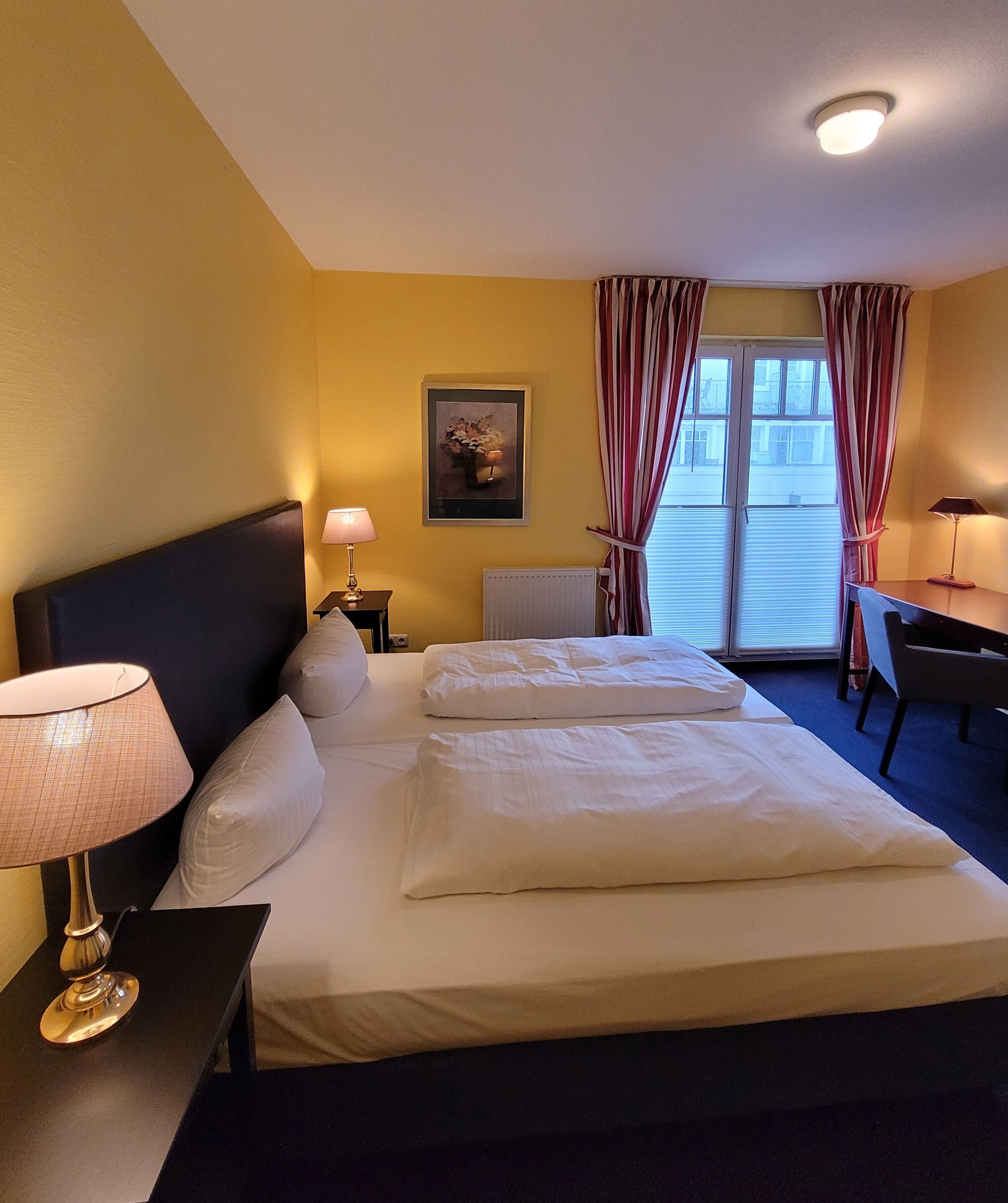 Quarto de hotel com uma cama grande, duas mesas de cabeceira, uma escrivaninha e uma janela com cortinas.