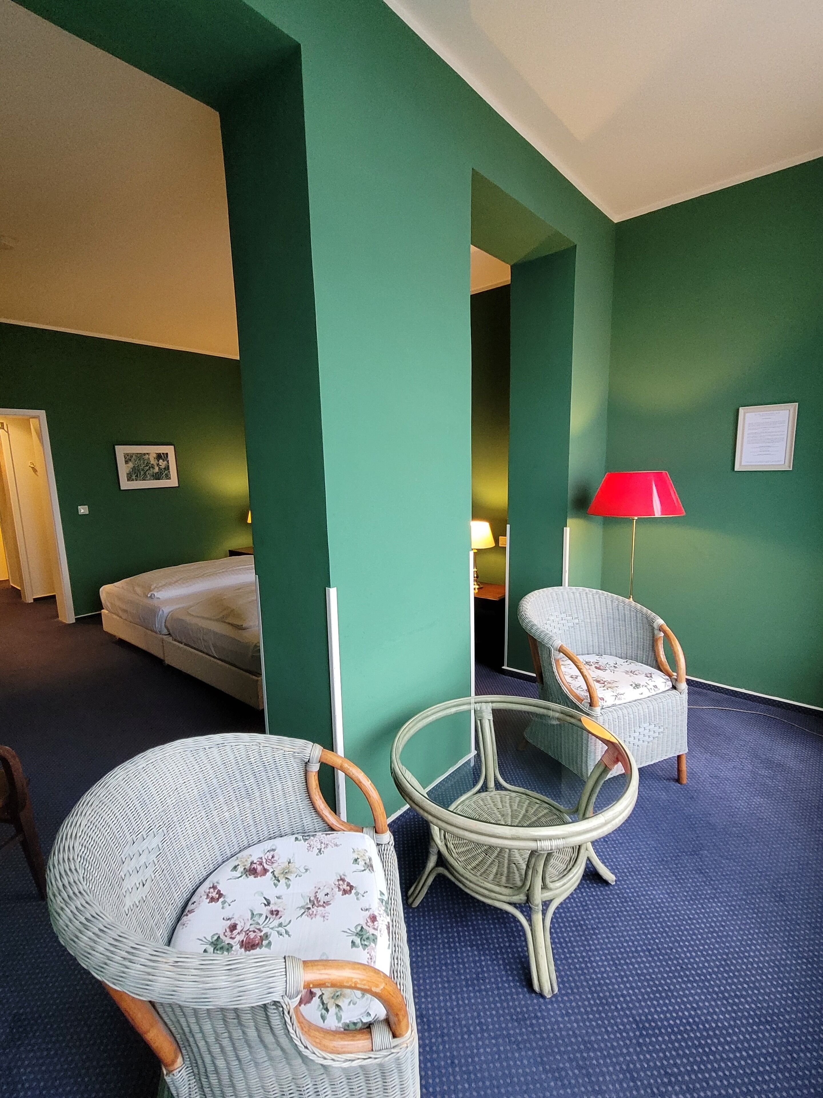 A room with green walls, a bed, two wicker chairs, a small table, and a red lamp.