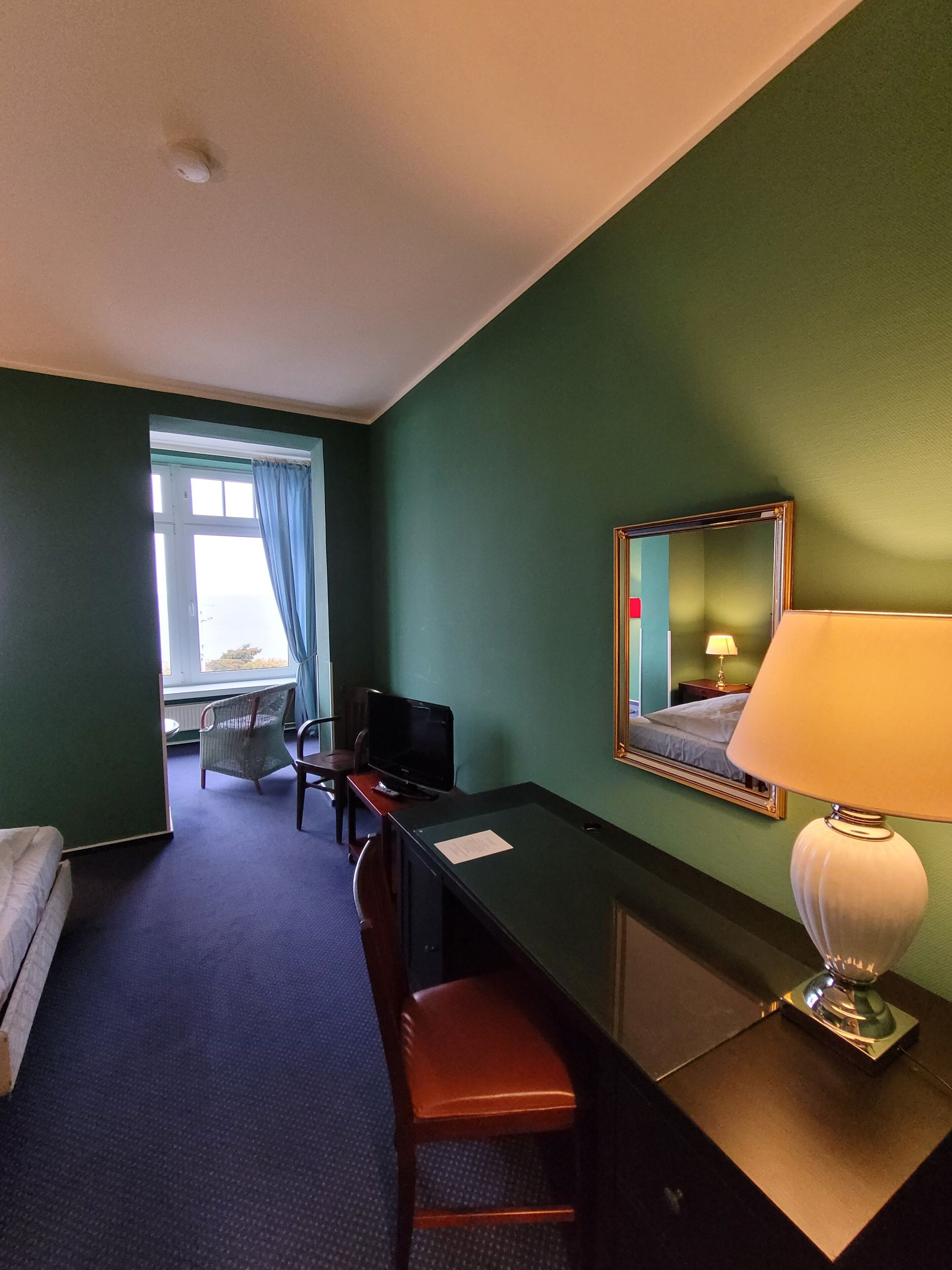 Et hotelværelse med en seng, et skrivebord med stol, en lampe, et spejl og et vindue med gardiner.